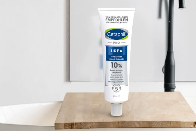 Das Cetaphil Fußpflegeprodukt "PRO Urea Aufbauende Intensiv-Fußsalbe" für extrem trockene und rissige Haut an den Füßen. Eine Tube stehend auf einem Holzregal im Badezimmer, neben einem Waschbecken und vor einem Spiegel.