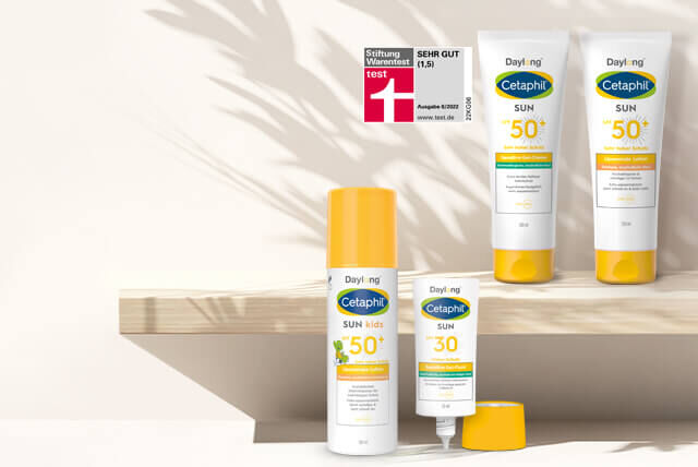 Vier Cetaphil SUN Sonnencremes für Kinder und Erwachsene vor weißer Wand mit leichtem Schatten einer Pflanze.